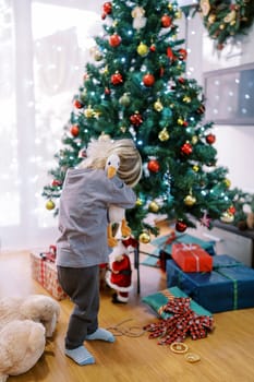 Little girl hugs a toy duck while standing near a decorated Christmas tree. Back view. High quality photo