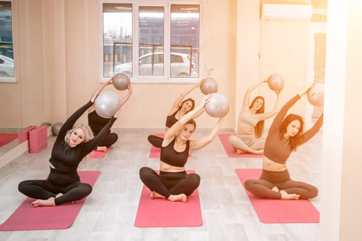 A group of six athletic women doing pilates or yoga on pink mats in front of a window in a beige loft studio interior. Teamwork, good mood and healthy lifestyle concept