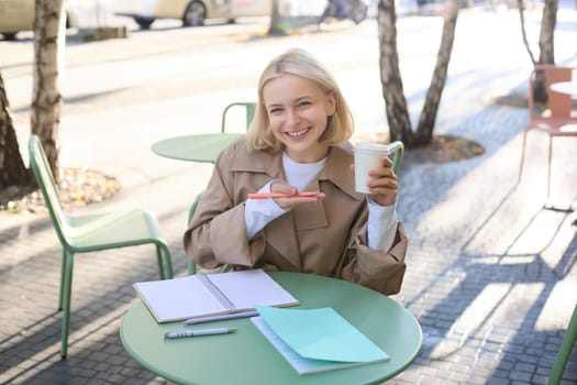 Happy, beautiful young european woman, laughing and smiling, holding coffee cup, working in outdoor cafe, writing in notebook, holding pen, looking at camera.