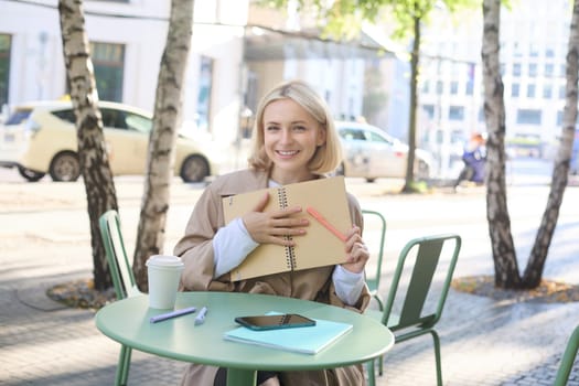 Image of young modern woman, student sitting in coffee shop with smartphone, holding notebook, showing her yellow journal, writing in planner and smiling.