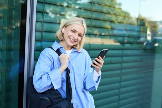 Portrait of beautiful blond girl, smiling woman in wireless earphones, listening to music on smartphone, waiting for friend on street, standing outdoors, leaning on building.