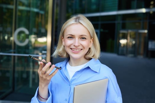 Close up portrait of smiling young female student, woman records voice message on smartphone, speaking on speakerphone while standing near office building, holding laptop.