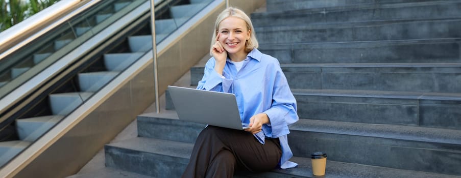 Image of stylish young woman, college student with laptop, sitting on city stairs and drinking coffee, working on project, studying outdoors, connects to public wifi, enjoying weather.