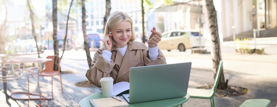 Cheerful young woman in trench coat with coffee, using wireless headphones and laptop, working remotely, connect to online class, video chatting.