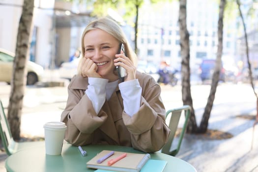 Close up of cheerful blond girl, woman talking on mobile phone, sitting in city centre, outdoor cafe, taking break from work or studying, chatting on telephone.