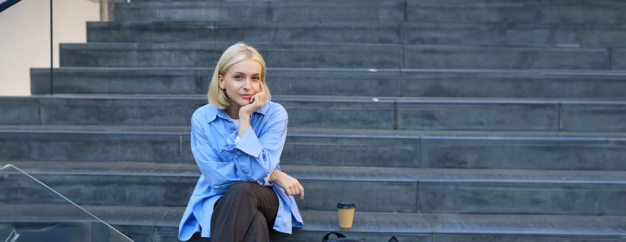 Urban style portrait of young woman, student sitting on city street stairs, with backpack, cup of coffee and laptop, looking happy and upbeat, smiling.