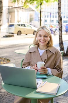 Vertical portrait of smiling blond woman, female student with coffee and laptop, has notebooks on table, studying remotely, working for online project, spending time outdoors on sunny day.