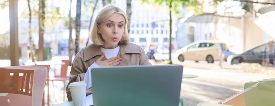 Close up portrait of young blond woman, sitting outdoors with laptop, drinking coffee in cafe, looking amazed and surprised at device screen, hears awesome news.