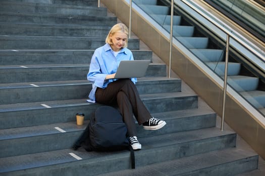 Portrait of woman with laptop, found wifi spot, sitting on stairs outdoors, drinking coffee, working on computer.