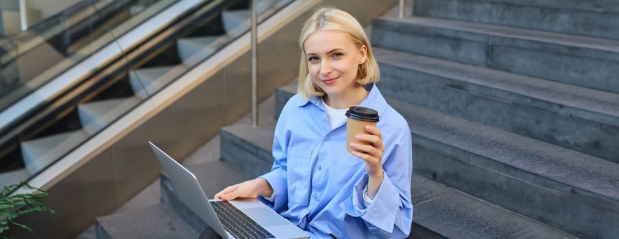 Image of stylish young modern woman, student doing homework, studying outdoors on campus stairs, sitting with laptop and coffee, drinking her cappuccino and connecting to public wifi.