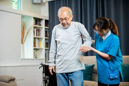 Asian doctor support old man to getting up to exercise, help handicapped elderly stand up, woman nurse assisting helping senior man patient get up from wheelchair for practice walking at home