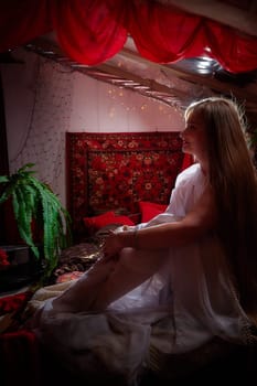 Beautiful European girl looking like Arab woman in red room with rich fabrics and carpets in sultan harem. Photo shoot of an oriental style odalisque. A model poses in sari as indian woman in india