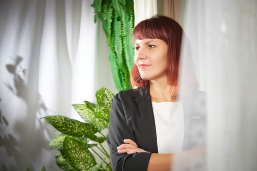 business woman in suit in an office or room standing by the window. The red-haired girl is ready to meet with clients and partners