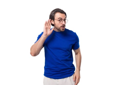 young smart brunette man with a beard dressed in a blue t-shirt overhears a secret on a white background.