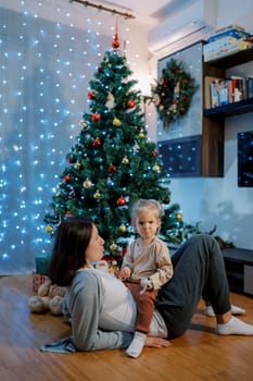 Little girl with pursed lips sits on her mom stomach lying on the floor near a decorated Christmas tree. High quality photo