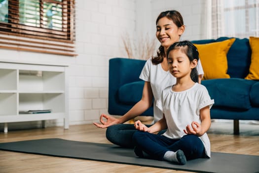 In a cozy home setting a young mother and her charming daughter practice family yoga in lotus position emphasizing mindfulness and meditation with smiles reflecting togetherness and happiness.