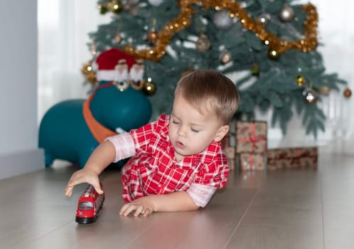 Christmas concept. New Year 2024. A small handsome boy in red checkered pajamas lies on the floor and plays with a red steam locomotive against the background of a decorated Christmas tree with gifts. Close-up.