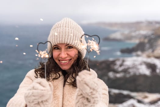 Outdoor winter portrait of elegant happy smiling beige hat, light faux fur coat holding heart sparkler, posing against sea and snow background