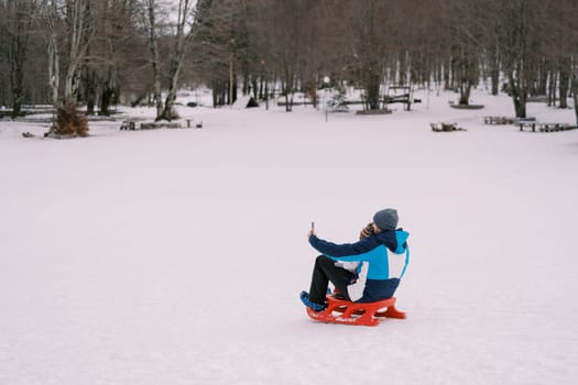 Dad taking smartphone selfie of himself and little kid sitting on sled on snowy lawn. Back view. High quality photo