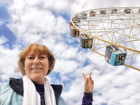 Relaxed mature caucasian female making selfie outside near big Ferris wheel. Positive senior woman against city street. Adult middle agged girl Blogger taking photo for internet