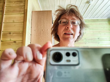 A shaggy, funny middle-aged woman in glasses takes selfie on her smartphone in front of the mirror in the morning. A blogger girl with disheveled hair takes pictures of herself for a blog