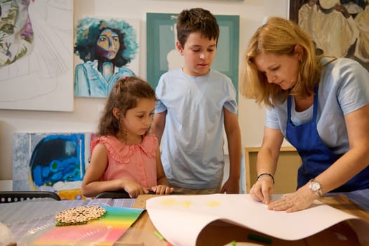 Caucasian charming female painter artist, instructor teaching assisting elementary age kids in painting at desk in art class. Kids development and entertainment. Fine art class. Education. People
