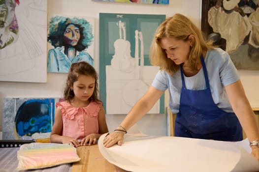 Confident young adult female art teacher rolling out a white paper sheet on the table, standing next to her little student girl in the creative art workshop. Artwork. Art gallery. Museum. Exhibition