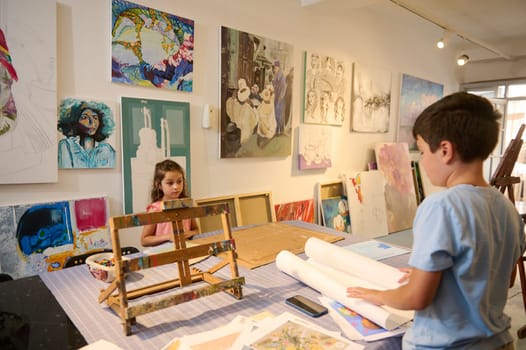 Adorable kids, a teenage boy and little child girl standing by a desk with pictures and drawing tools in the art class, learning art. Fine art, visual art, kids entertainment and development concept