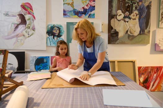 Authentic portrait of a little Caucasian cute child girl in casual pink dress, and her inspired female teacher at art class, standing against the background of painted pictures displayed on the wall