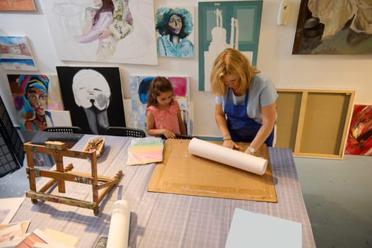 View from above to a Caucasian little child girl student learning art in creative art class. Female artist teaching painting to an elementary age student, a gallery of paints on the wall on background