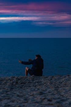 Man who is sitting at the edge of the beach next to water having videocall and showing beautiful cloudy sunset at golden hour at the beach next to dark and cold sea at summer right after storm