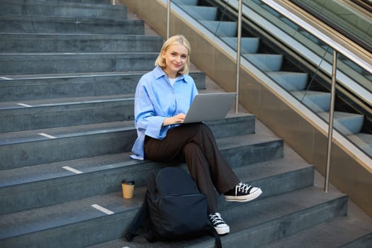 Portrait of young woman, student sitting outdoors on stairs with coffee, using laptop, working on project, looking at camera and smiling.