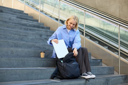 Image of stylish young woman, putting away her laptop in backpack, sitting on stairs near campus, packing for university, drinking coffee.