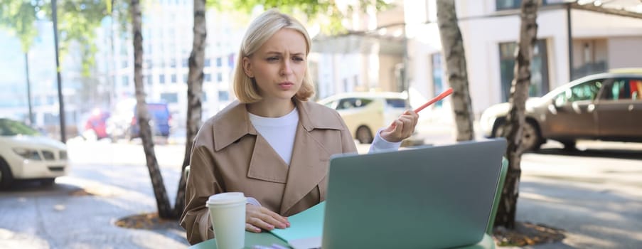 Image of woman with confused face, looking at laptop, working remotely, chatting with someone and shrugging, talking with puzzled face, sitting in coffee shop.