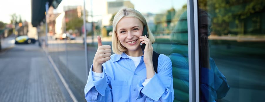 Close up portrait of blond smiling woman with backpack, standing on street near office building, showing thumb up in approval, recommending smth good.