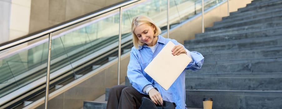 Image of stylish young woman, putting away her laptop in backpack, sitting on stairs near campus, packing for university, drinking coffee.