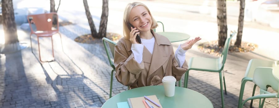 Outgoing young smiling woman, sitting in outdoor cafe, drinking coffee, answer phone call, talking on mobile, chatting with friendly attitude on smartphone.