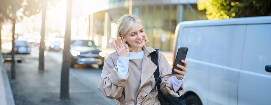 Portrait of happy young woman walking on street, waving hello at smartphone camera, connects to online video chat, calling someone on mobile phone on her way to work.