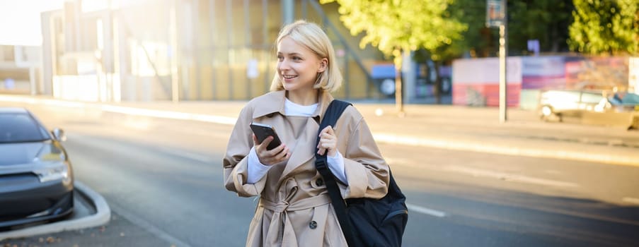 Modern young woman with backpack, student using smartphone, standing on street, sunny day, waiting for ride, using mobile phone application.