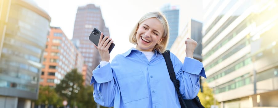 Portrait of happy, enthusiastic young woman, holding backpack and mobile phone, standing on street and rejoicing, triumphing, making fist pump gestures, dancing from happiness and joy.