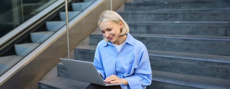 Smiling girl student using laptop computer modern technology device outdoor in university campus online learning, elearning outside sitting on urban stairs. Web education course webinars concept.