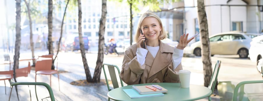 Image of stylish blond woman talking on mobile phone, drinking coffee in cafe outdoors, enjoying warm weather in city centre, answer telephone call on smartphone.