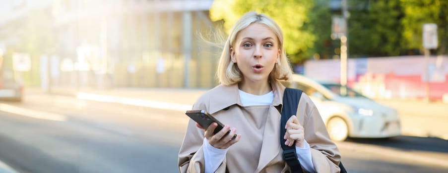 Image of young surprised woman, standing on street with mobile phone, looking amazed at camera, saying wow.