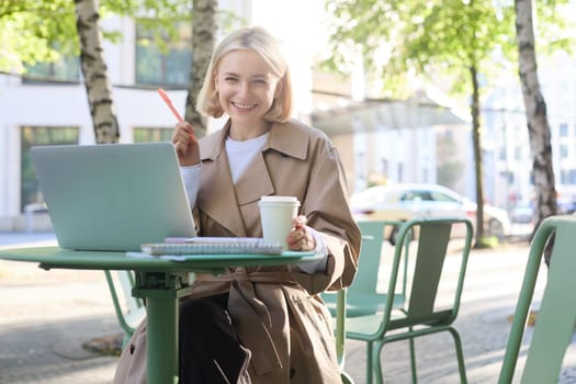 Portrait of young smiling blond woman, working on laptop, sitting in outdoor cafe on street, drinking coffee. Lifestyle and people concept