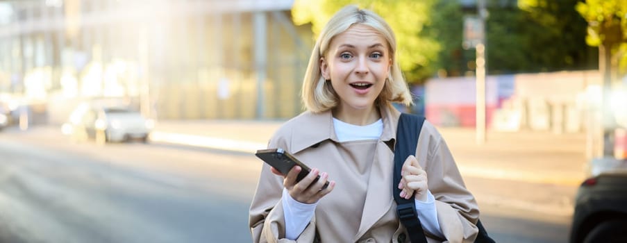 Portrait of young blond woman with backpack, holding mobile phone, standing on street on sunny day, looking surprised and amazed at camera, expresses interest, hear great news.
