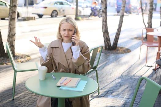 Image of upset, confused young woman, talking on mobile phone and shrugging, sitting in cafe, struggle to understand something over the telephone.