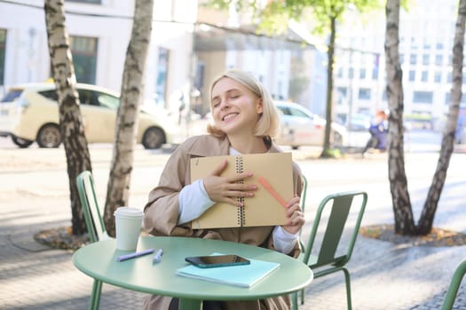 Image of young beautiful woman, student doing homework in an outdoor coffee shop, holding journal or planner, writing in notebook, smiling happily.