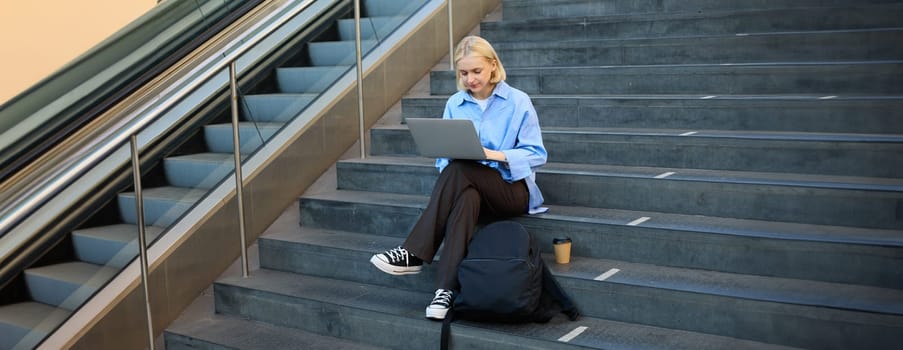 Portrait of woman with laptop, found wifi spot, sitting on stairs outdoors, drinking coffee, working on computer.