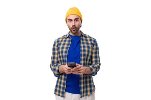 young stylish emotional brunette man with a brutal beard and mustache in a hat and shirt looks at the camera in surprise holding the phone in his hand.