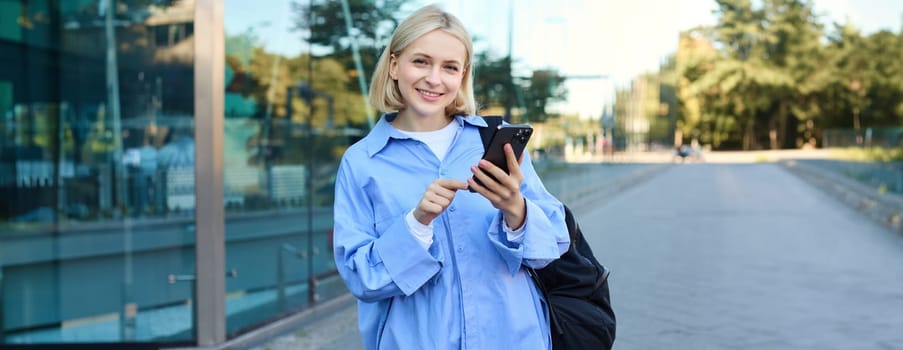 Portrait of smiling, modern blond woman walking along the street in blue collar shirt, has backpack on shoulder, messaging, using mobile phone app.